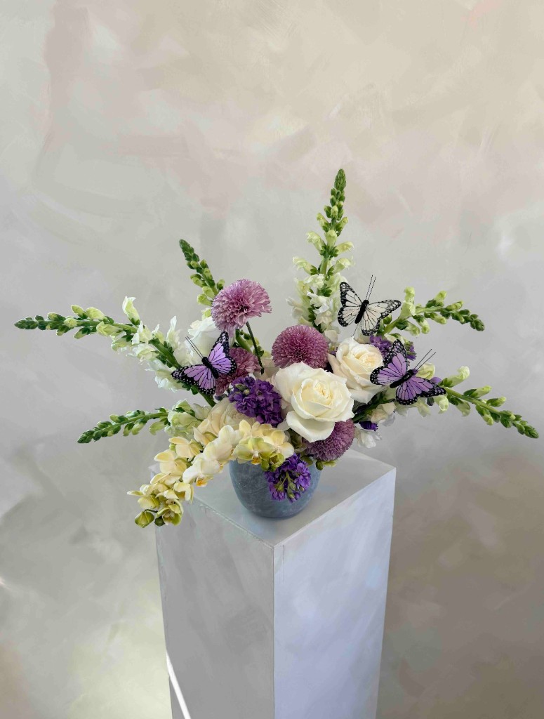 Elegant floral arrangement with white roses, purple pompon flowers, and decorative butterflies in a blue-gray vase on a white pedestal