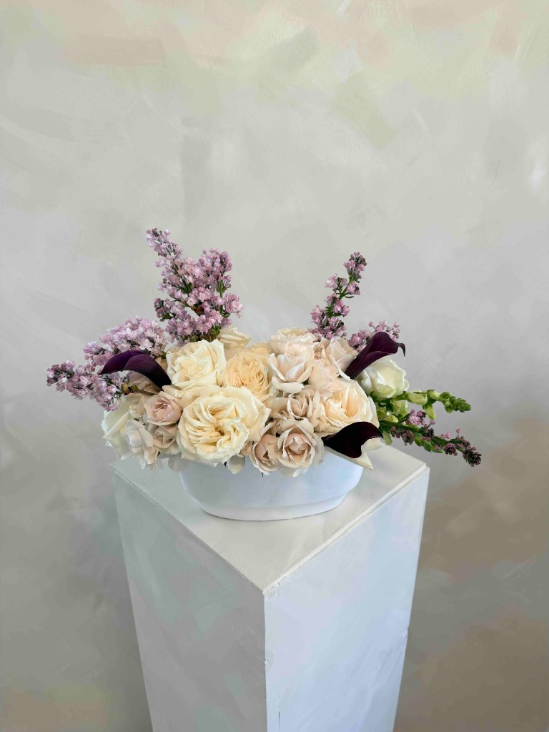 Low white arrangement with cream roses, blush blooms, lavender accents, and deep plum calla lilies on a white pedestal