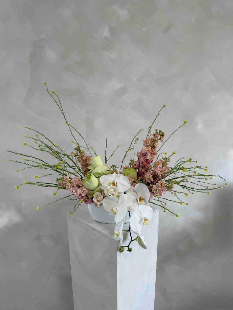 White Phalaenopsis orchids, mint-green roses, dusty pink stock, and branches with yellow buds in a pale blue bowl on a gray pedestal