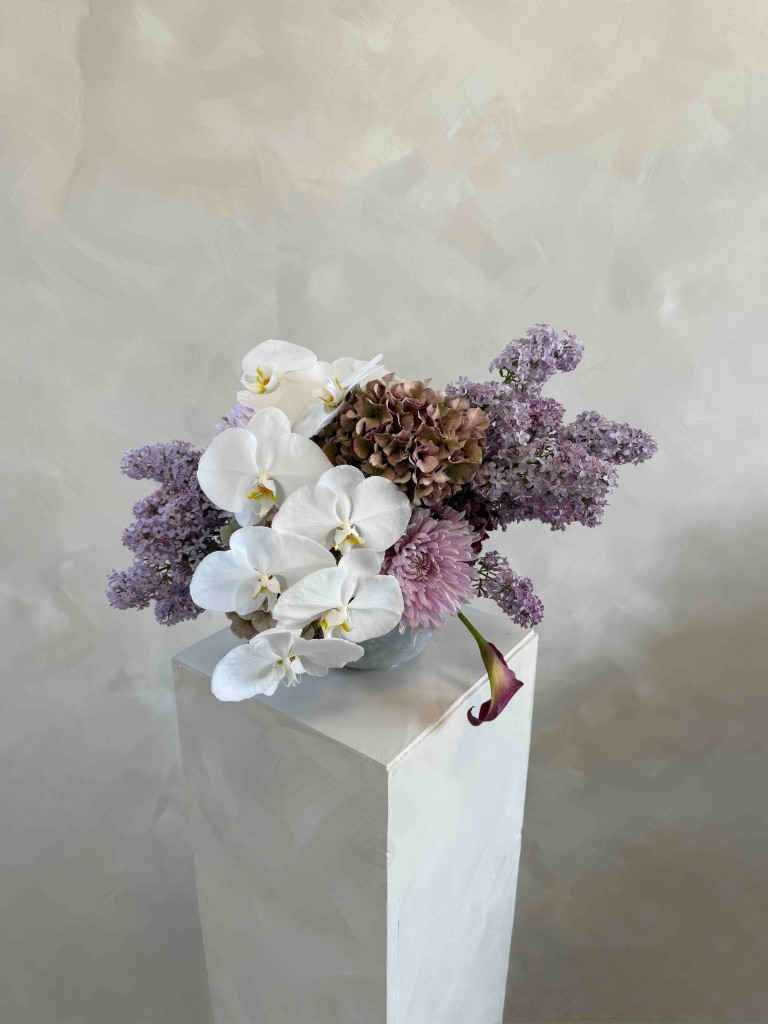 Studio arrangement with white orchids, lilac, bronze hydrangea, mauve blooms, and a plum calla lily in a pale blue-gray bowl on a white pedestal
