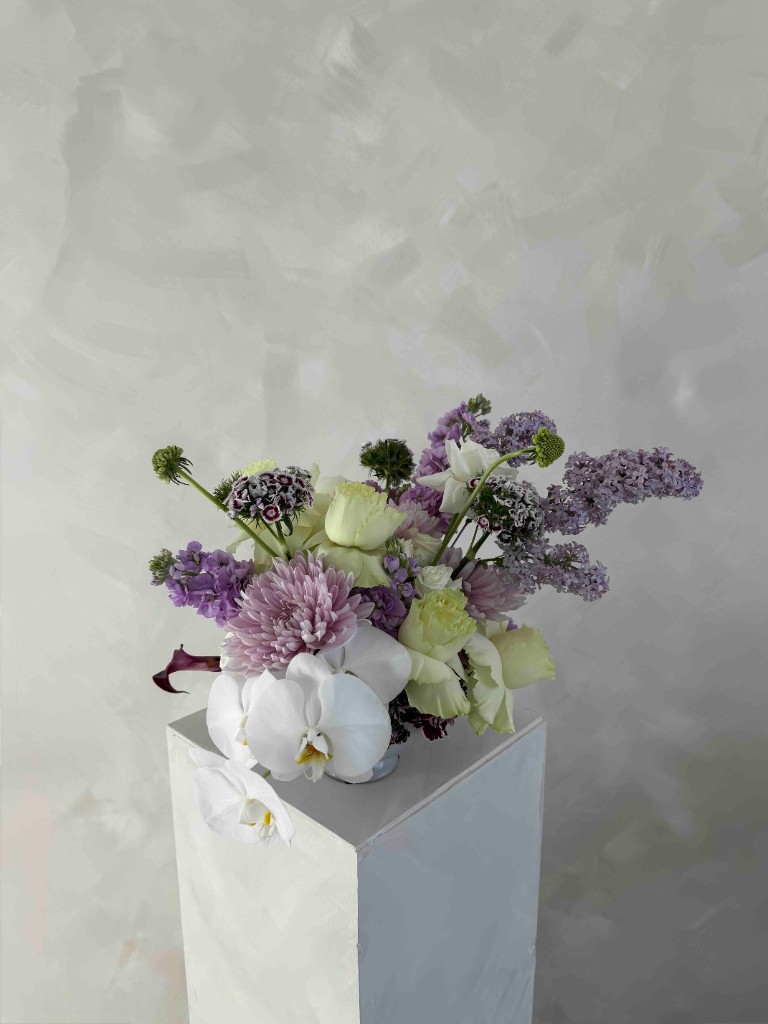 Romantic arrangement with white orchids, cream roses, mauve chrysanthemums, lavender lilacs, and plum accents on a white textured pedestal