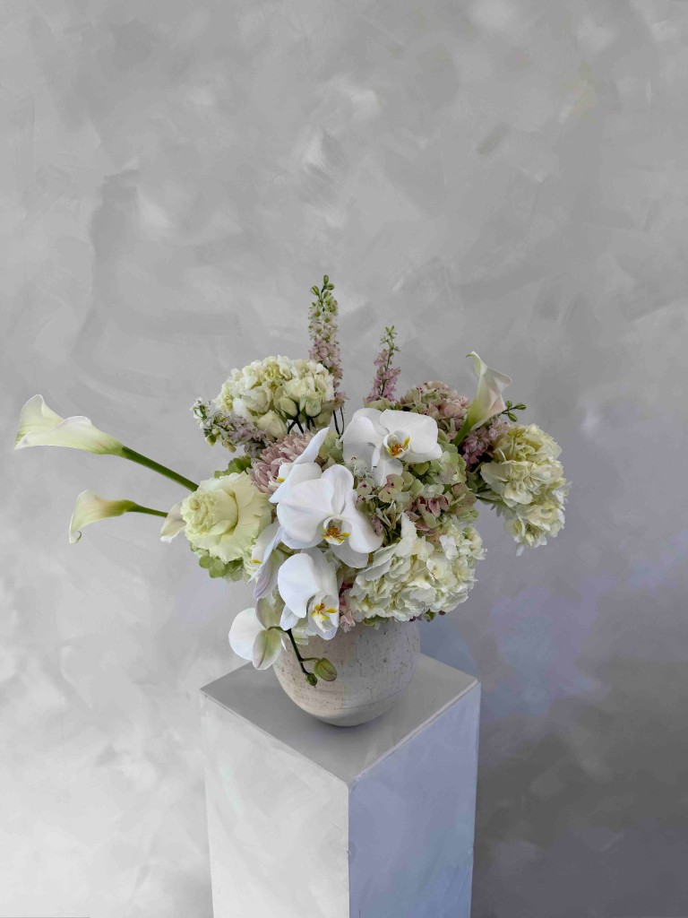 Muted arrangement with white orchids, hydrangeas, calla lilies, pale pink vertical blooms, and green accents in a textured beige vase on a white pedestal