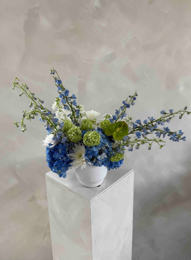 Closer view of an airy arrangement with blue hydrangeas, pale delphinium, white mums, lime green accents, and a green anthurium in a white vase on a pedestal