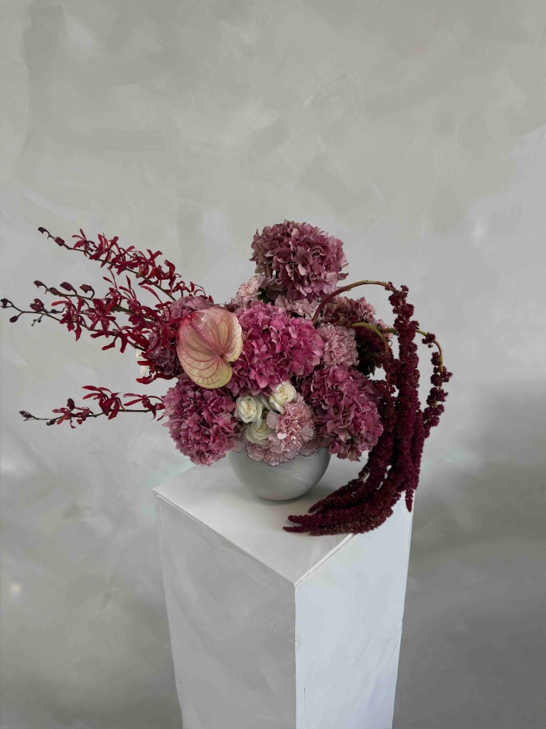 Dramatic arrangement with dusty pink hydrangeas, burgundy orchids, trailing amaranthus, cream roses, and a pale pink anthurium in a light grey bowl on a white pedestal