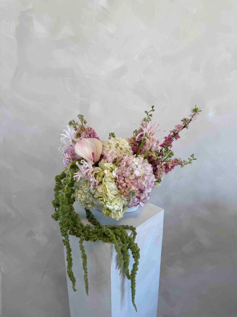 Pastel arrangement with pink and cream hydrangeas, a pink anthurium, branching pink blooms, and cascading green amaranthus in a white bowl on a pedestal