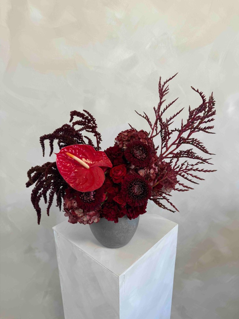 Moody burgundy and crimson arrangement with a red anthurium, dark roses, trailing amaranth, dusty pink hydrangea, and sculptural branches in a grey bowl on a white pedestal