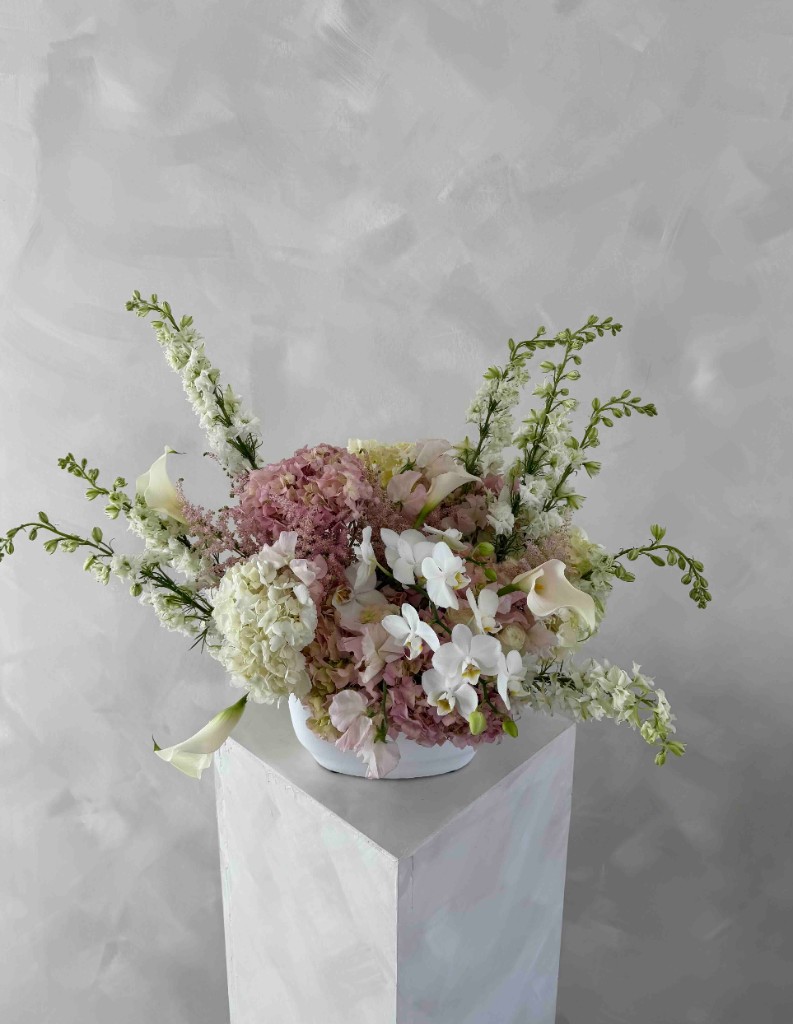 Lush pink and white hydrangeas, orchids, calla lilies, and tall white spikes in a low white bowl on a white square pedestal against a textured gray wall
