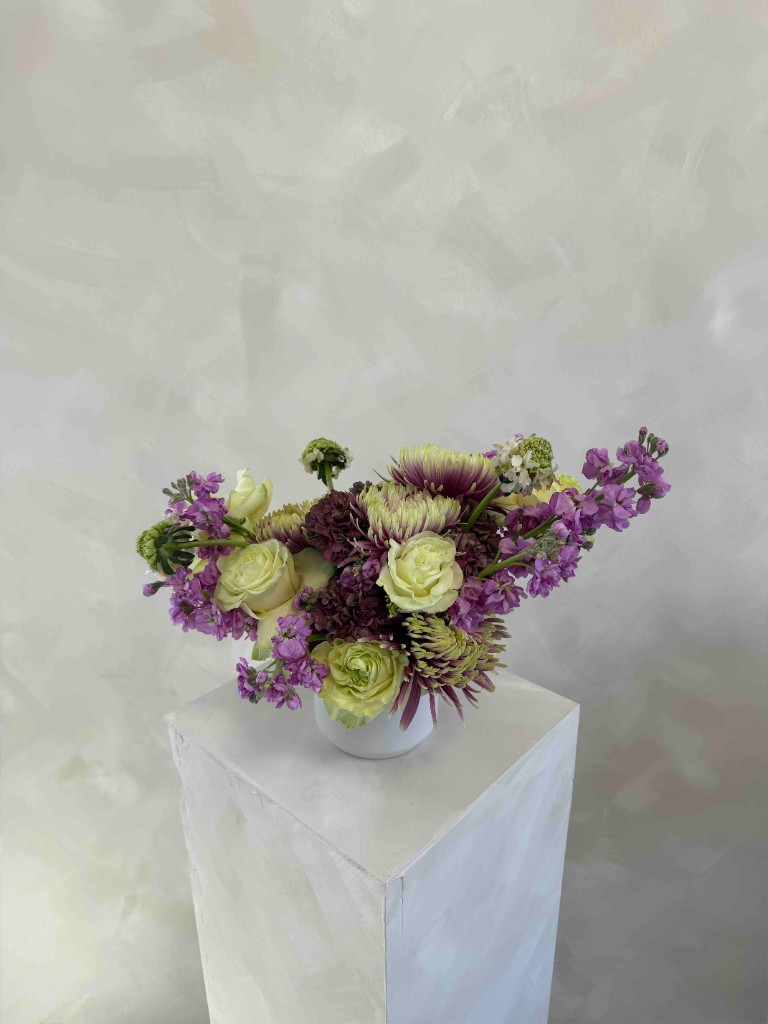 Low white arrangement with lavender stock, green and cream roses, mauve chrysanthemums, and purple accents on a white pedestal against a textured gray wall