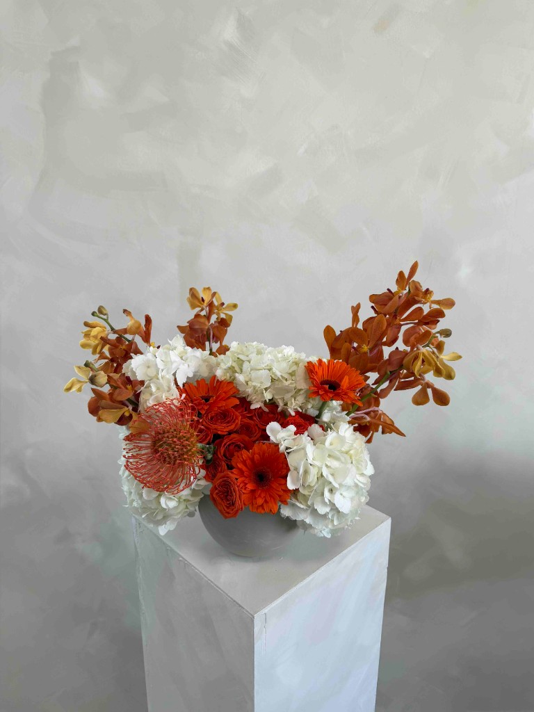 Orange and white arrangement with orchids, hydrangeas, gerberas, and roses in a low white bowl on a pedestal