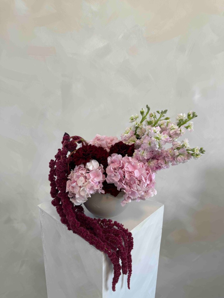 Pink hydrangea arrangement with deep burgundy blooms and cascading amaranthus in a white bowl on a pedestal