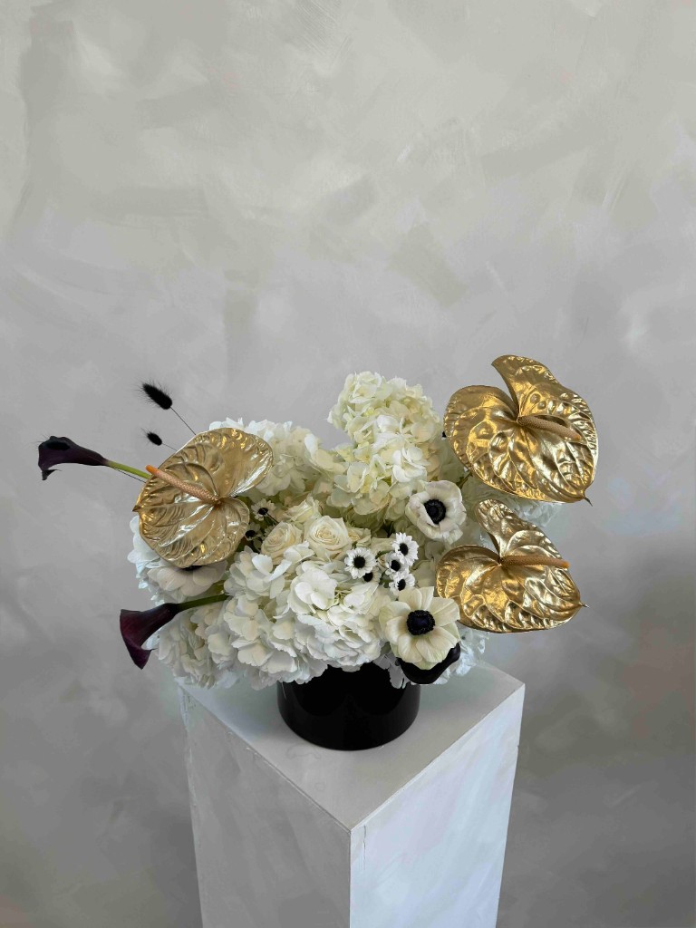 White arrangement with hydrangeas, calla lilies, anemones, and gold anthuriums in a black vase on a pedestal