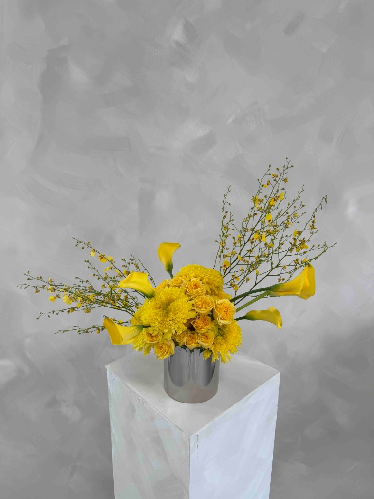 Vibrant yellow arrangement with chrysanthemums, roses, calla lilies, and dancing lady orchids in a metallic silver vase on a white pedestal