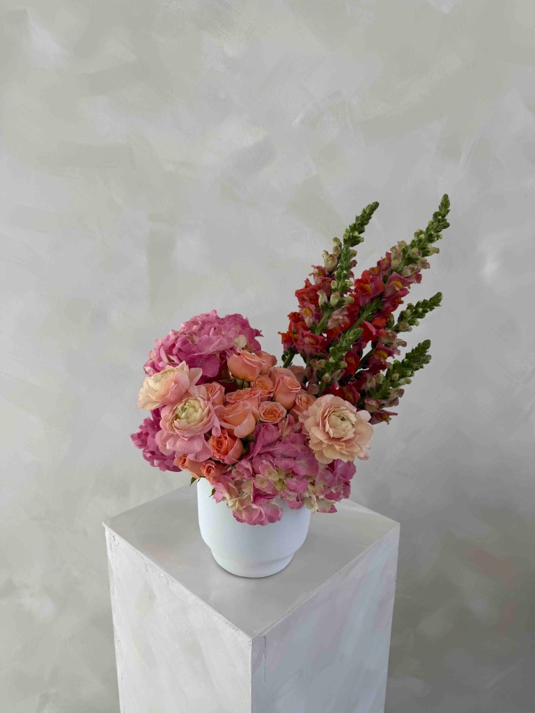 Lush arrangement with pink hydrangeas, peach ranunculus, coral roses, and tall pink snapdragons in a white vase on a pedestal against a textured grey wall
