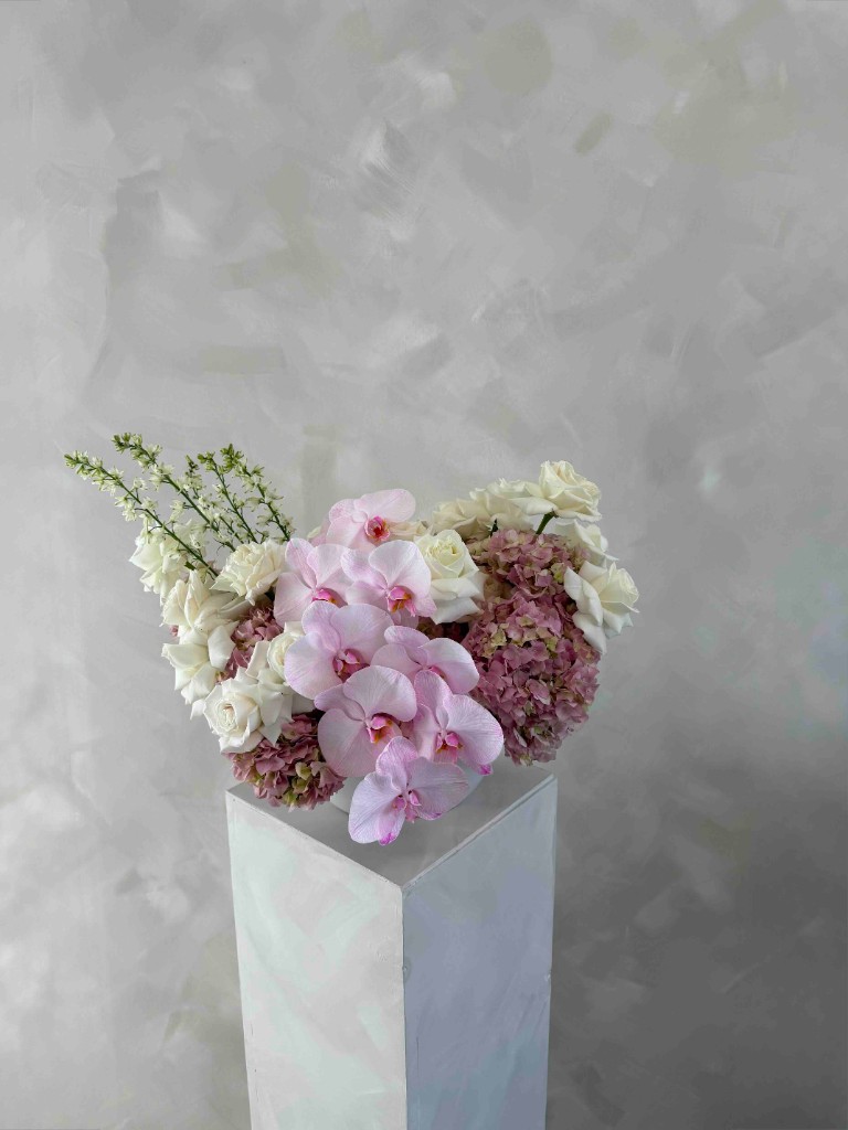 Sophisticated arrangement with pink Phalaenopsis orchids, creamy white roses, mauve hydrangeas, and tall white flower spikes in a white bowl on a pedestal