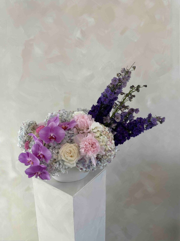 Elegant arrangement with magenta orchids, soft pink blooms, tall indigo delphiniums, and pale blue hydrangeas in a white bowl on a pedestal