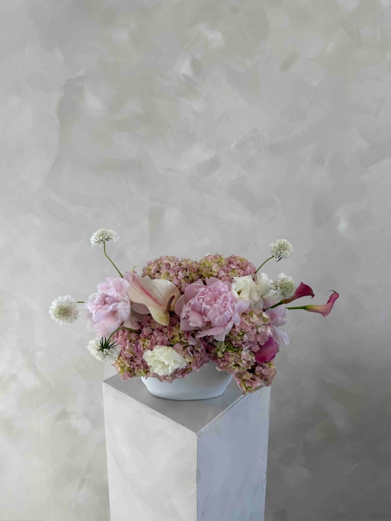 Romantic centerpiece with pink peonies, hydrangeas, pale and deep pink calla lilies, and airy white accents in a white oval bowl on a pedestal