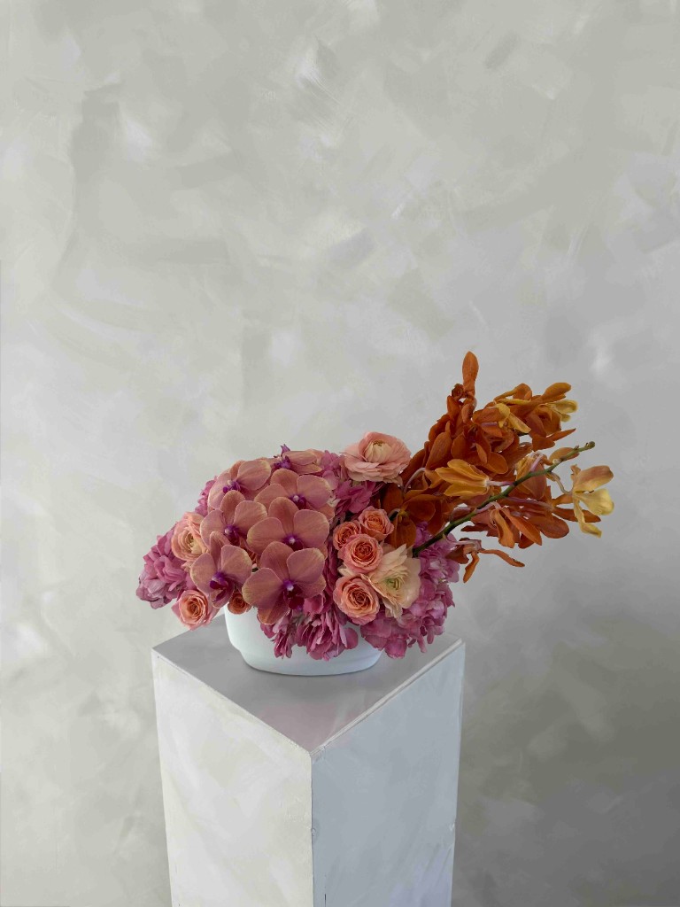 Warm-toned arrangement with peach and pink orchids, spray roses, ranunculus, rust-orange orchid spikes, and pink hydrangeas in a white bowl on a pedestal
