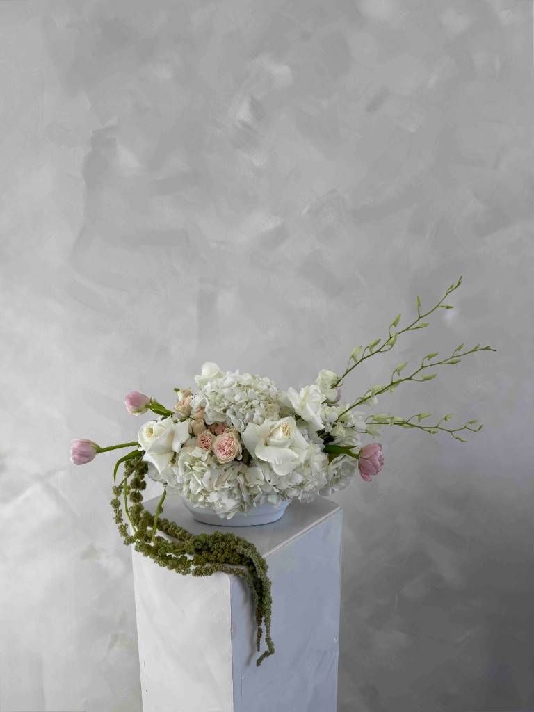 Airy arrangement with white hydrangeas and roses, soft pink roses and tulips, trailing green vines, and budding stems in a white bowl on a pedestal