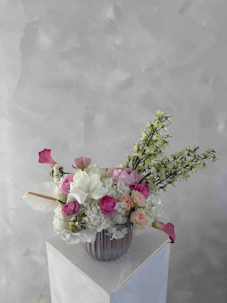 Lush pink and white arrangement with ranunculus, roses, hydrangeas, calla lilies, a white anthurium, and pale vertical blooms in a ribbed lavender vase on a pedestal