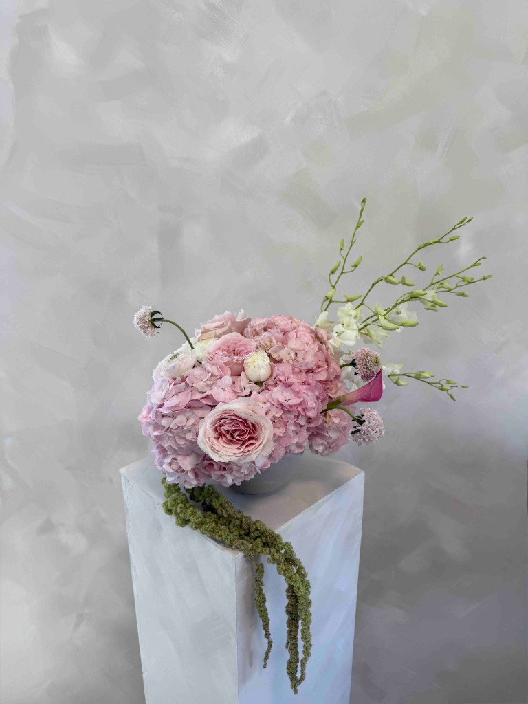 Pink hydrangeas and roses with white accents and trailing green amaranthus in a grey bowl on a white pedestal against a textured grey wall