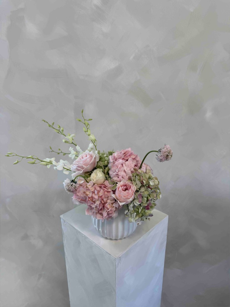 Pastel arrangement with pale pink roses, pink and sage hydrangeas, and white orchid-like blooms in a ribbed white vase on a pedestal