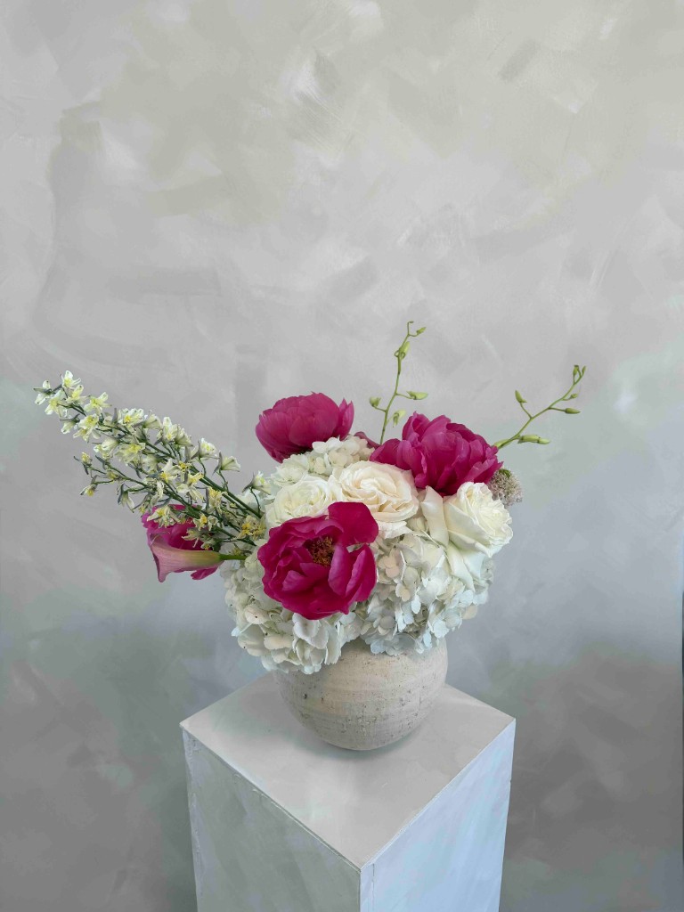 Vibrant magenta peonies with white hydrangeas and roses in a textured ceramic vase on a white pedestal