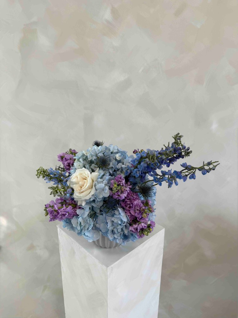 Cool-toned arrangement with light blue hydrangeas, purple stock, a creamy white rose, and blue delphinium in a ribbed vase on a white pedestal