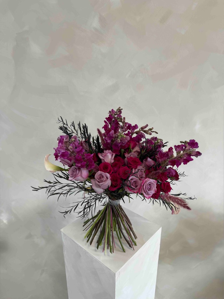 Bouquet of lavender and deep red roses with magenta snapdragons, a creamy calla lily, and dark feathery foliage on a white pedestal