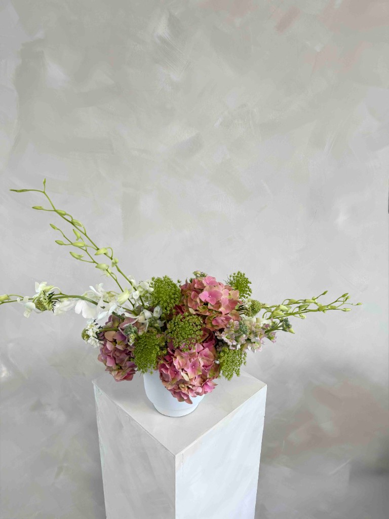 Pink hydrangeas, green filler, and white orchids in a white vase on a white pedestal against a textured gray wall