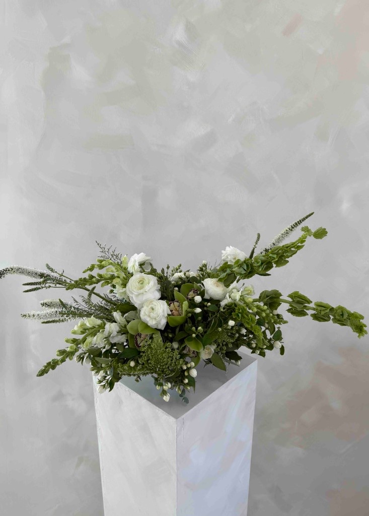 White and green asymmetrical arrangement with ranunculus, veronica, green orchids, and bells of Ireland on a white pedestal against a textured gray wall