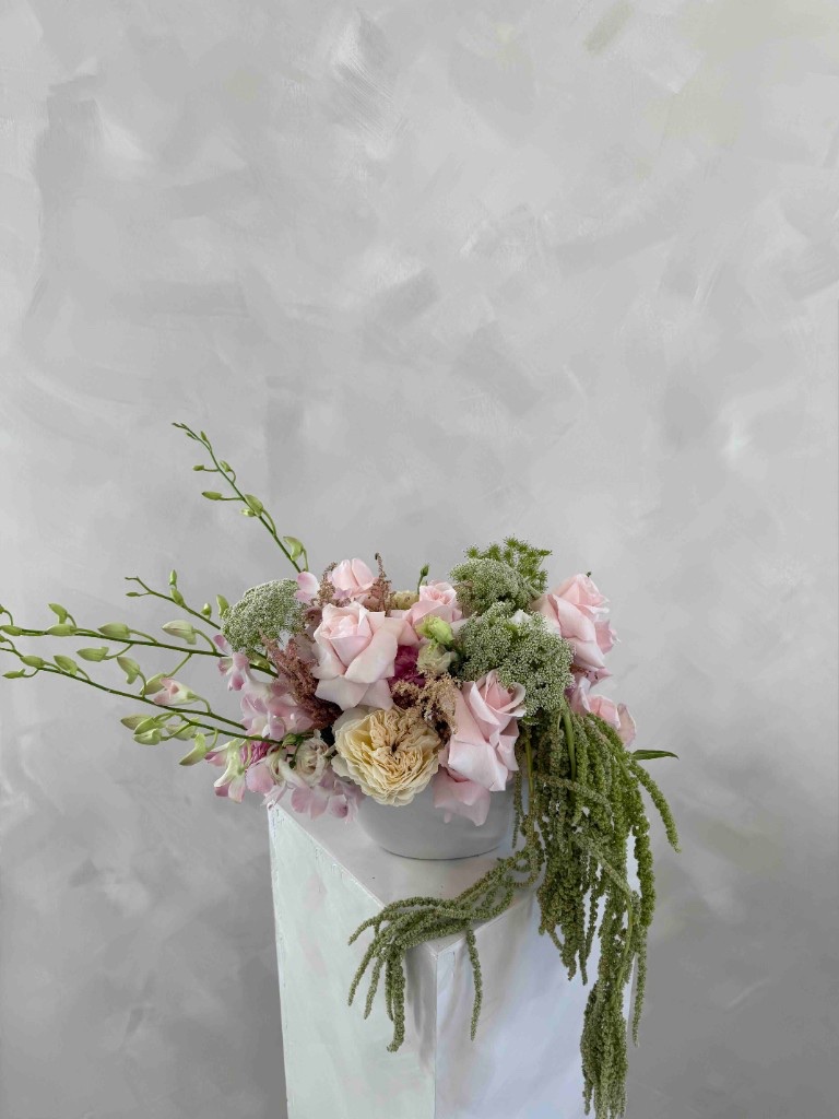 Pastel pink and cream arrangement with roses, hydrangeas, Queen Anne's lace, and trailing green amaranth in a white bowl on a pedestal against a textured gray wall