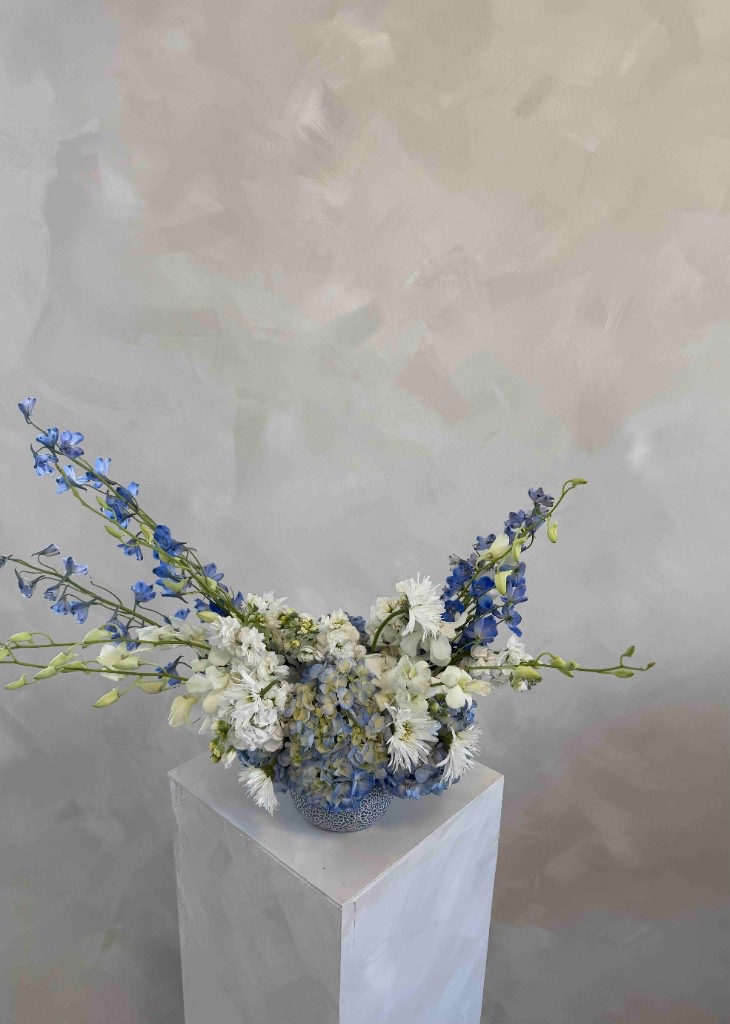 Blue and white wing-shaped arrangement with hydrangeas, delphinium, and pale green buds in a speckled blue and white vase on a white pedestal