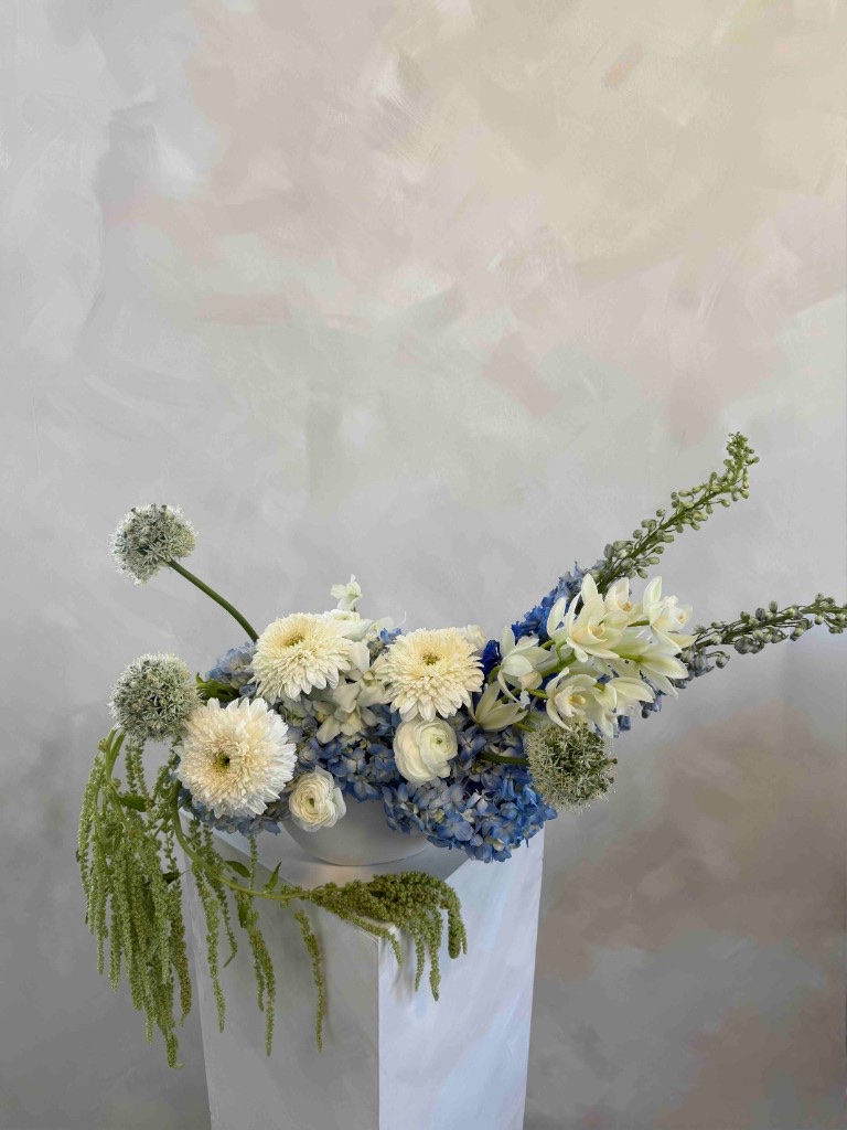 Cool-toned arrangement with white chrysanthemums, blue hydrangeas, white orchids, and green allium in a white bowl on a pedestal with trailing green amaranth