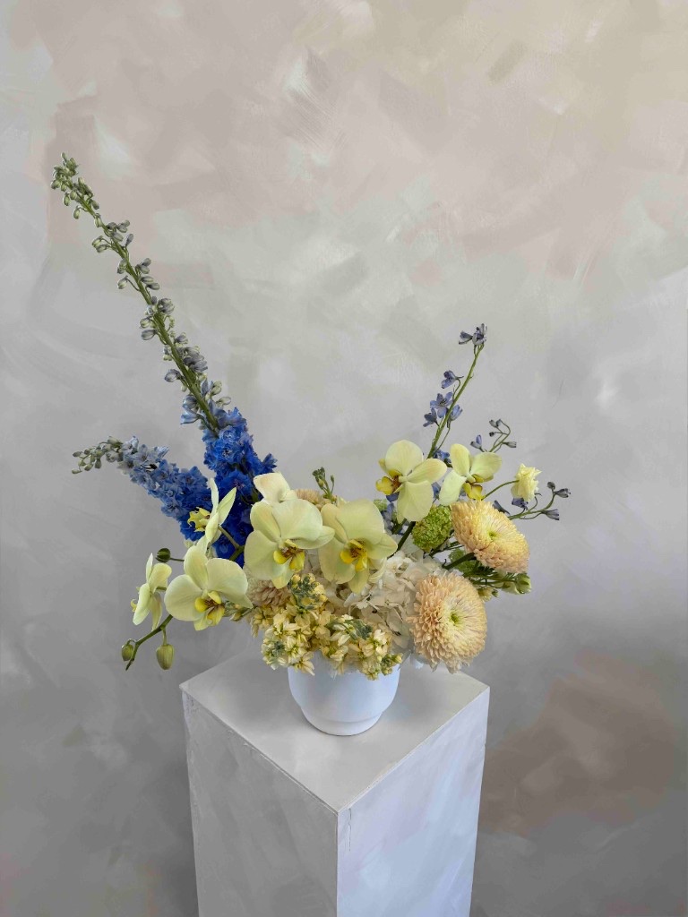 Asymmetrical arrangement with blue delphinium, yellow-green orchids, peach pompon chrysanthemums, and pale yellow stock in a white bowl on a pedestal