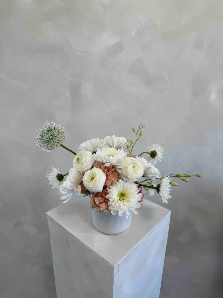 Delicate white ranunculus and chrysanthemums with peach carnations and a white allium in a white cylindrical vase on a pedestal against a textured gray wall