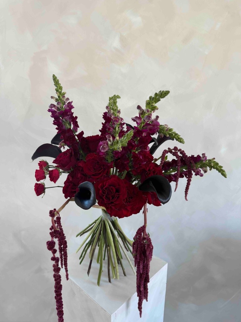 Moody hand-tied bouquet with deep red roses, dark purple snapdragons, black calla lilies, and burgundy amaranthus on a white pedestal