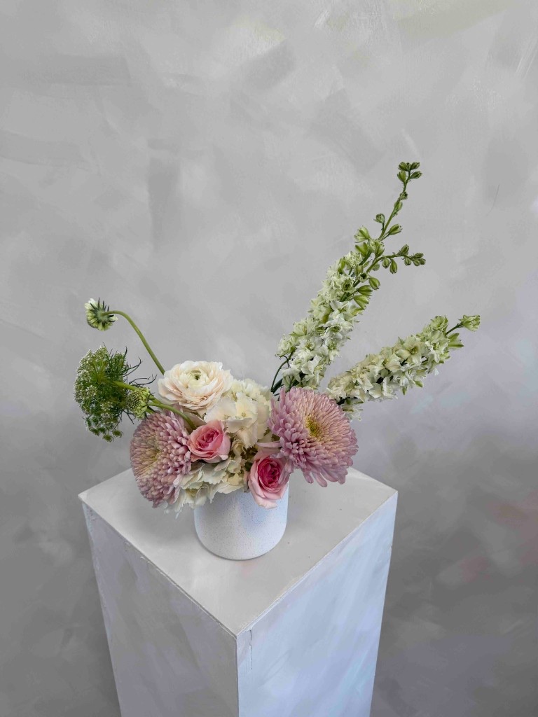 Airy arrangement with lavender chrysanthemums, pink roses, white ranunculus, white hydrangeas, and white delphinium in a speckled white vase on a pedestal