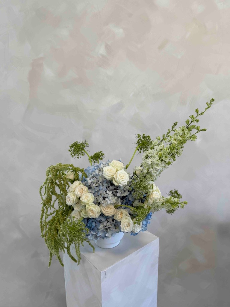 Asymmetrical arrangement with pale blue hydrangeas, white roses, green larkspur, Queen Anne's lace, and trailing green amaranthus in a white bowl on a pedestal