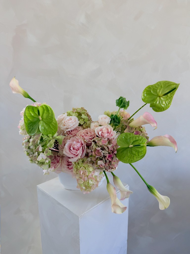 Arrangement with pink and cream roses, hydrangeas, anthurium leaves and white calla lilies in a round white vase