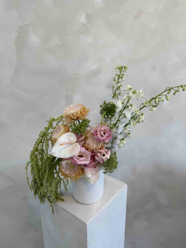 Pastel peach and blush arrangement with chrysanthemums, lisianthus, white anthurium, white delphinium, and trailing green amaranthus in a white vase on a pedestal