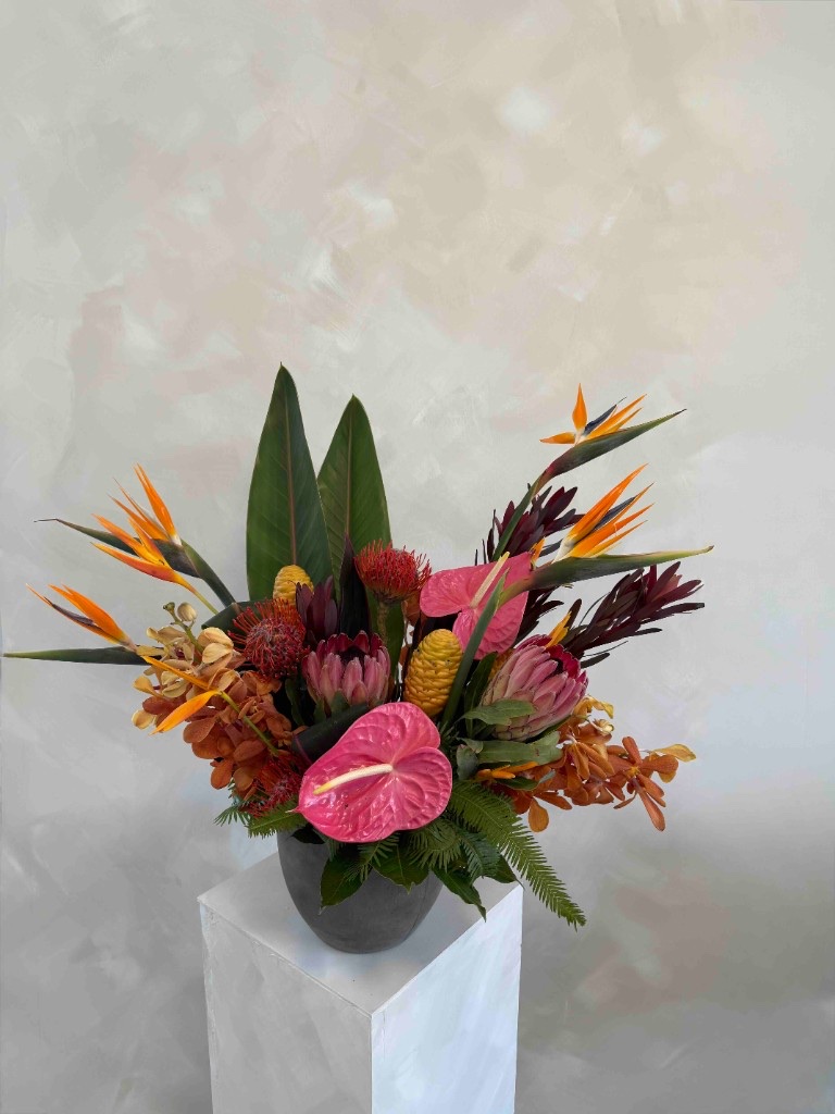 Vibrant tropical arrangement with pink anthuriums, birds of paradise, proteas, beehive ginger, and orchids in a dark grey pot on a white pedestal