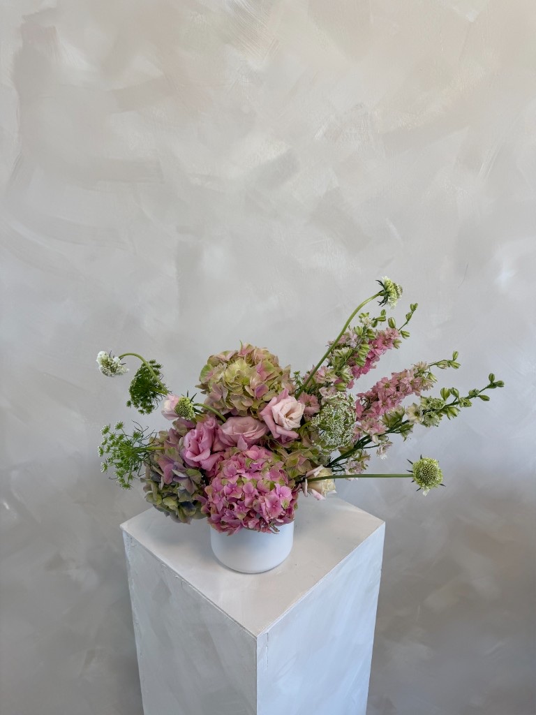 Romantic arrangement with dusty pink and green hydrangeas, mauve roses, pink delphinium, and Queen Anne's lace in a white vase on a pedestal
