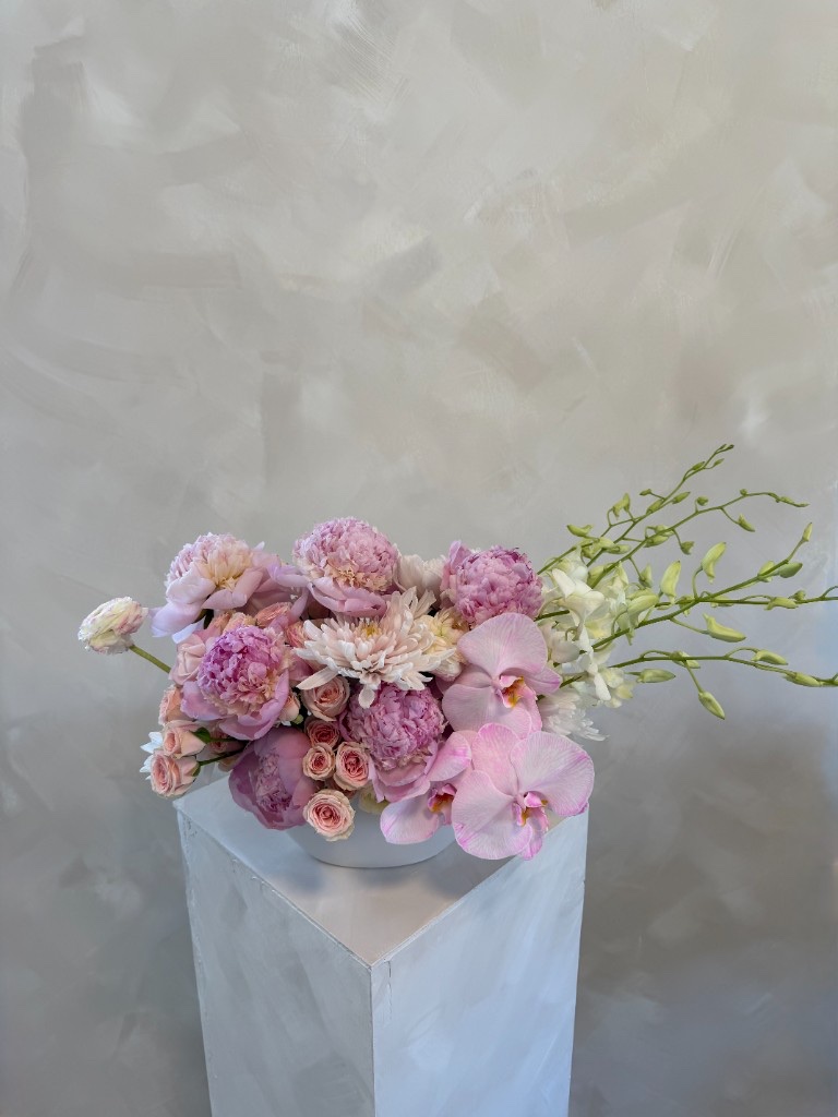 Lush pink arrangement with peonies, orchids, spray roses, and a white chrysanthemum in a white bowl on a pedestal against a textured gray wall