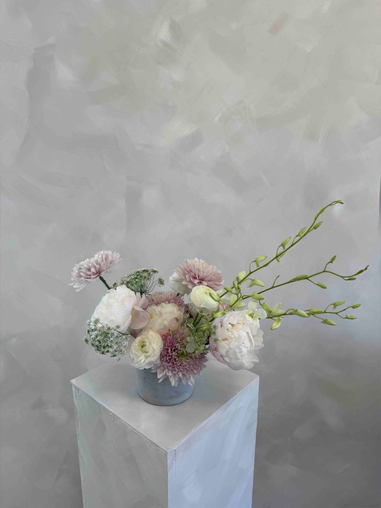 Pastel arrangement with white peonies, blush chrysanthemums, white ranunculus, and Queen Anne's lace in a blue-gray vase on a white pedestal