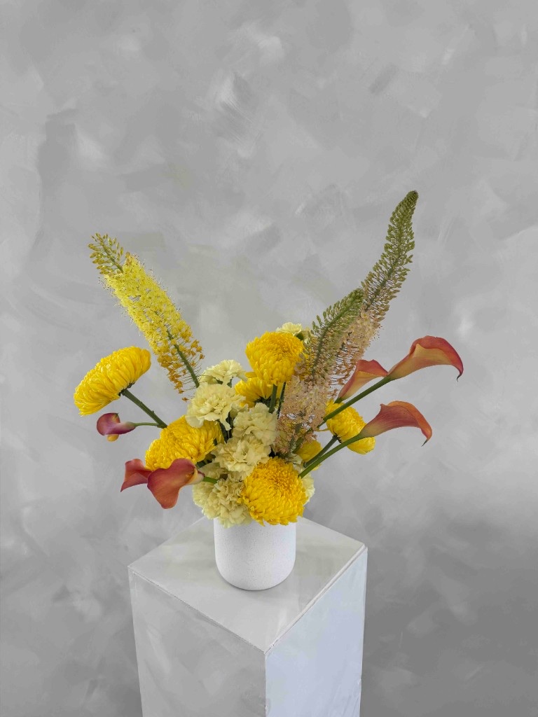 Vibrant yellow and orange arrangement with chrysanthemums, calla lilies, and foxtail lilies in a white vase on a pedestal against a textured gray wall
