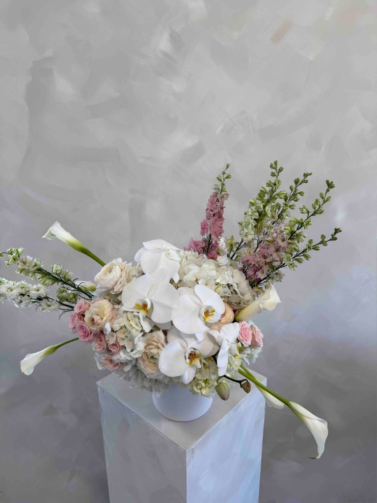 Asymmetrical white and pink arrangement with Phalaenopsis orchids, hydrangeas, ranunculus, calla lilies, and pink larkspur in a white bowl on a pedestal