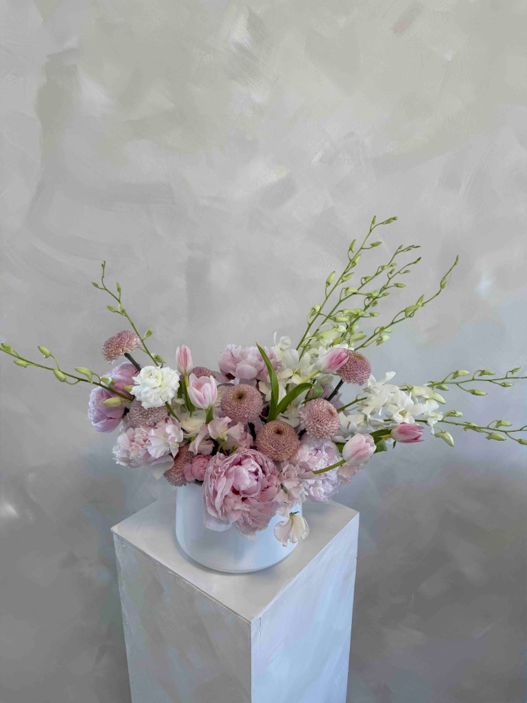 Pastel pink and white arrangement with peonies, tulips, pompon dahlias, and airy green orchid stems in a white vase on a pedestal