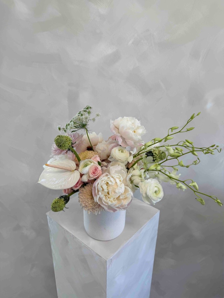 Sophisticated pastel arrangement with white peonies, white anthurium, pale pink roses, ranunculus, and Queen Anne's lace in a white vase on a pedestal