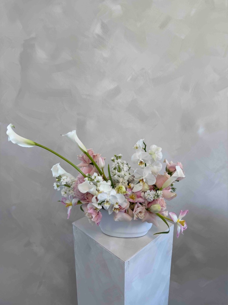 Asymmetrical white and pink arrangement with calla lilies, orchids, roses, and stock in a white bowl on a pedestal against a textured gray wall