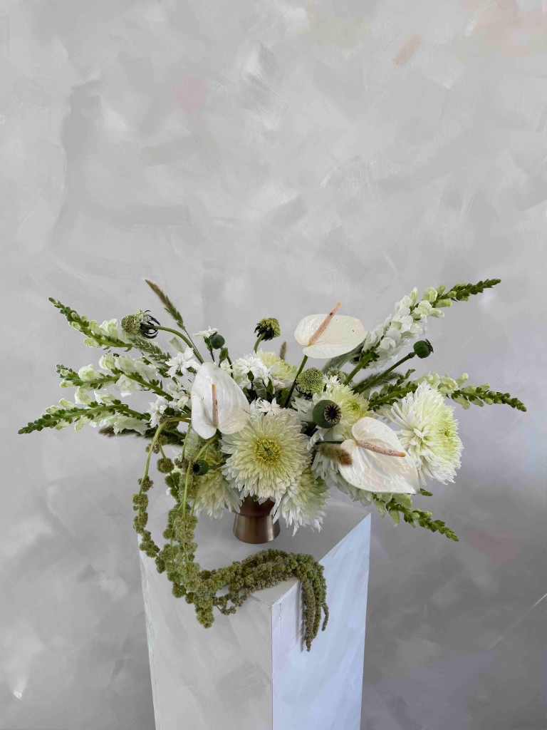 White and green arrangement with chrysanthemums, anthuriums, snapdragons, poppy seed pods, and trailing green amaranthus in a bronze vase on a white pedestal